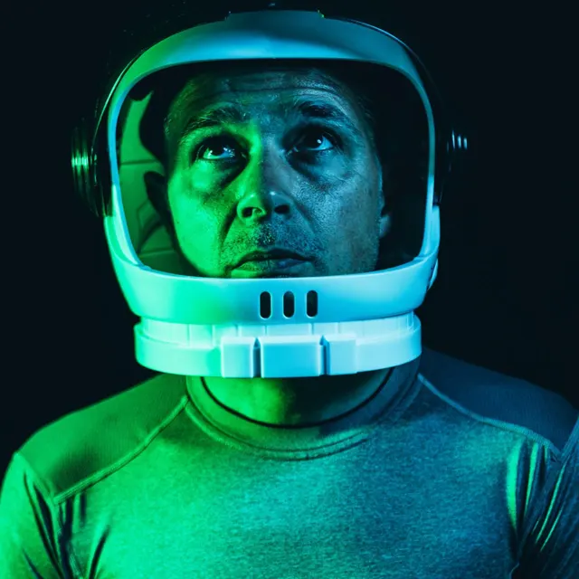 A man in an astronaut helmet lit in green, looking up